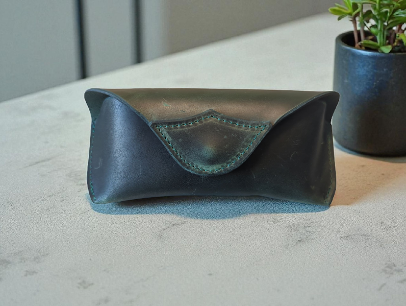 Dark Green leather eyeglass case on a light gray surface with a plant in the background